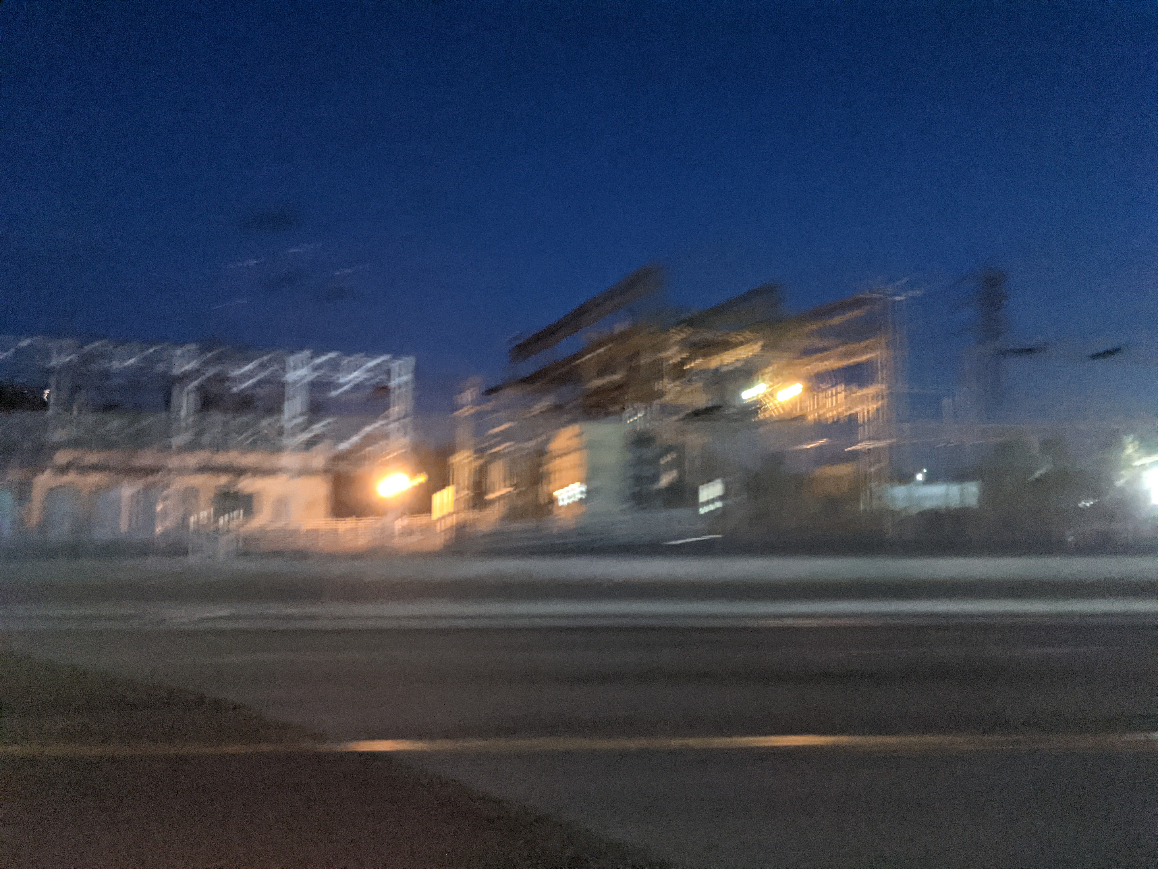 A motion blurred photo of a brightly lit roadside power station under the clear evening sky, with the shadow of a car partially visible on the road.
